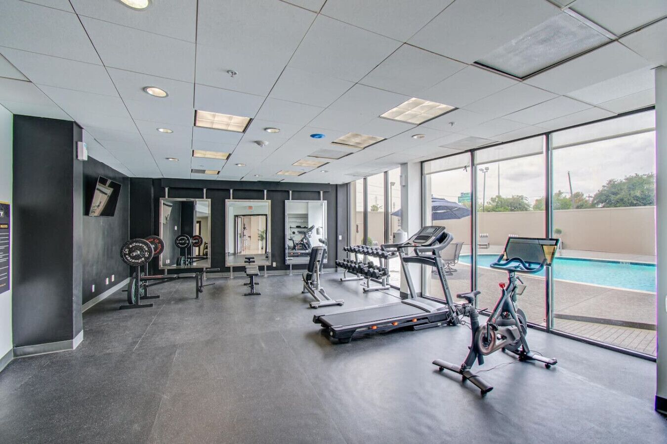 A gym with cardio machines, weightlifting equipment, and a large window overlooking a swimming pool.