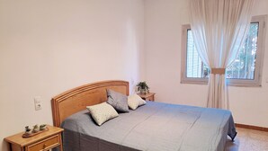Quarto com cama, mesa de cabeceira, janela com cortinas e pequena mesa com plantas.