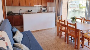 Cozinha compacta com armários de madeira, área de jantar com mesa e cadeiras, e varanda com vista para edifícios.