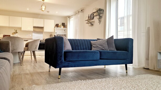 Un salon moderne avec un canapé bleu, un canapé gris et un tapis blanc. On aperçoit au fond une cuisine avec des meubles blancs et un coin repas.