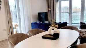 Un salon moderne avec une table de salle à manger ronde, des chaises recouvertes d’un tissu beige, un canapé bleu et un téléviseur.