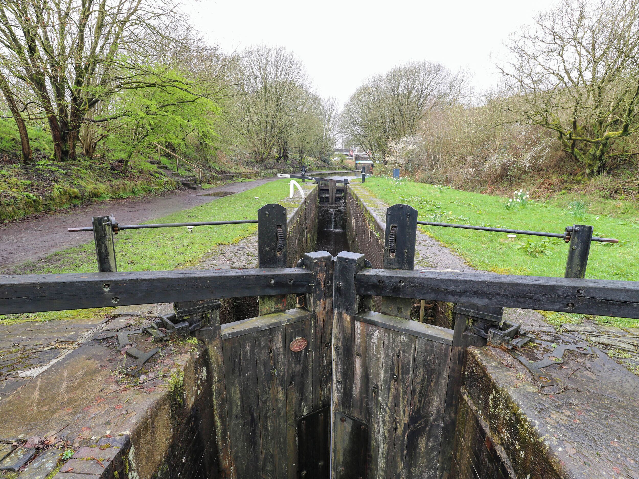 A wooden lock gate with a metal mechanism, surrounded by greenery and a pathway.