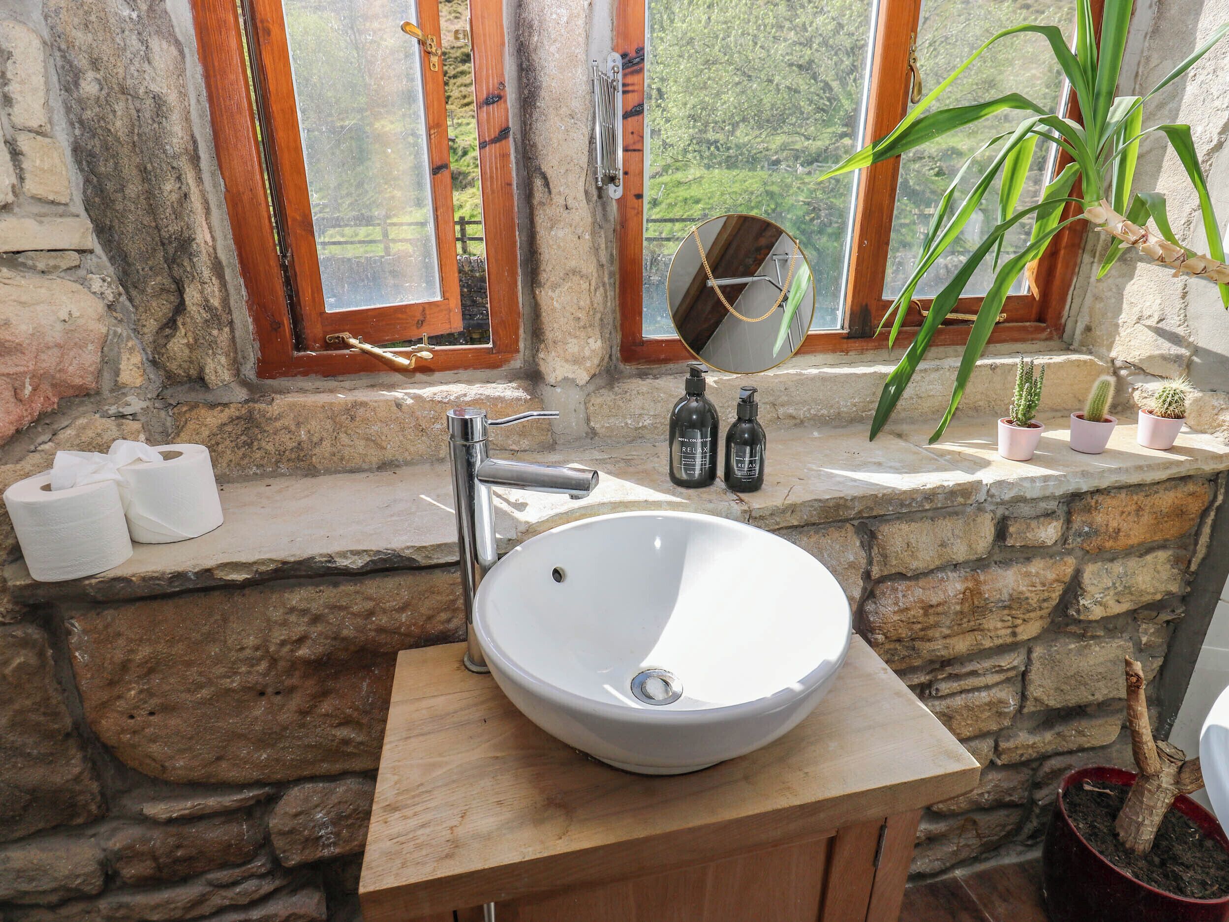 A bathroom with a stone wall, wooden vanity, a white sink, and a window with a view of greenery.