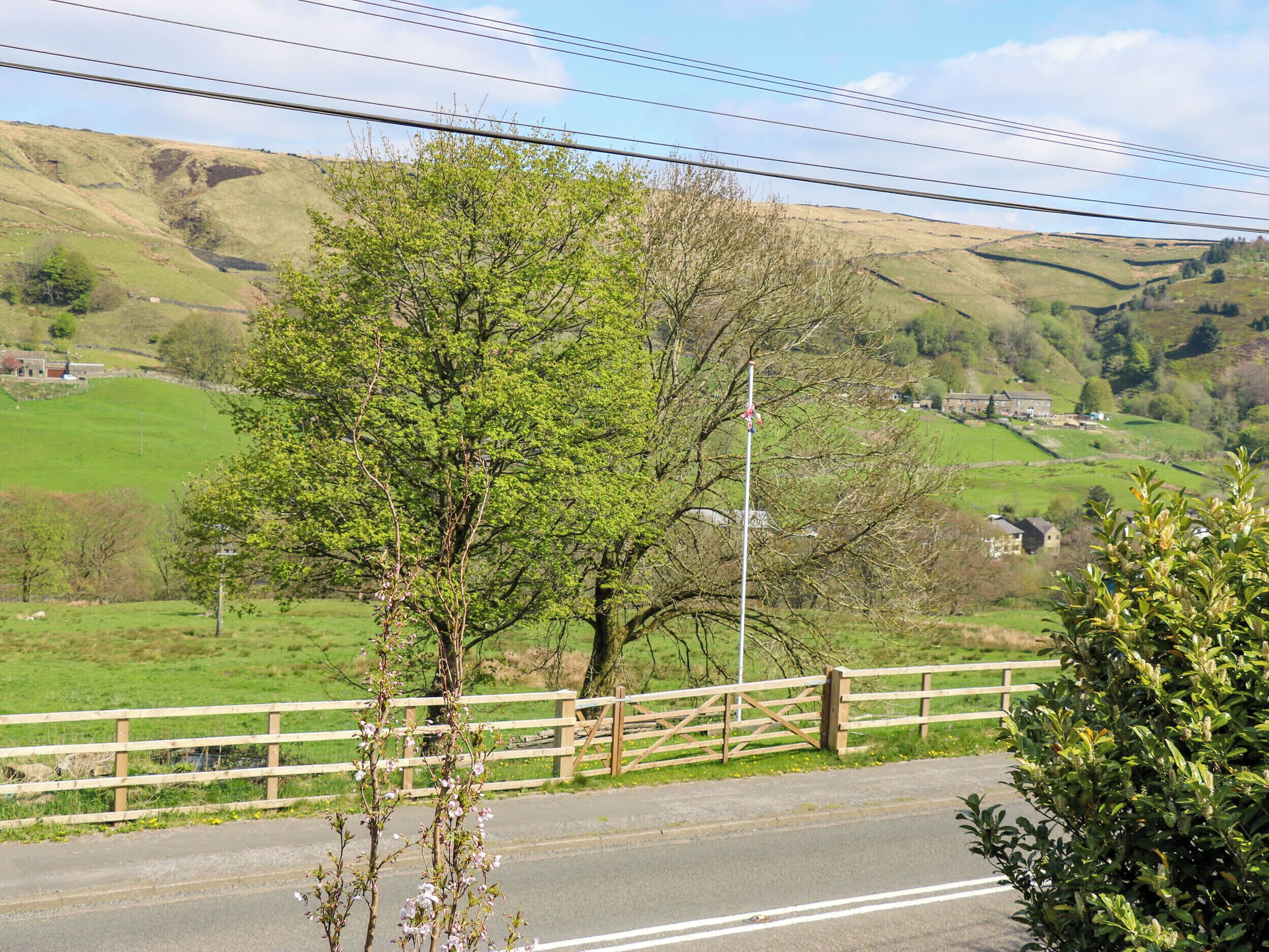 Un camino rural con una valla de madera, un árbol y una vista distante de una casa en una colina.