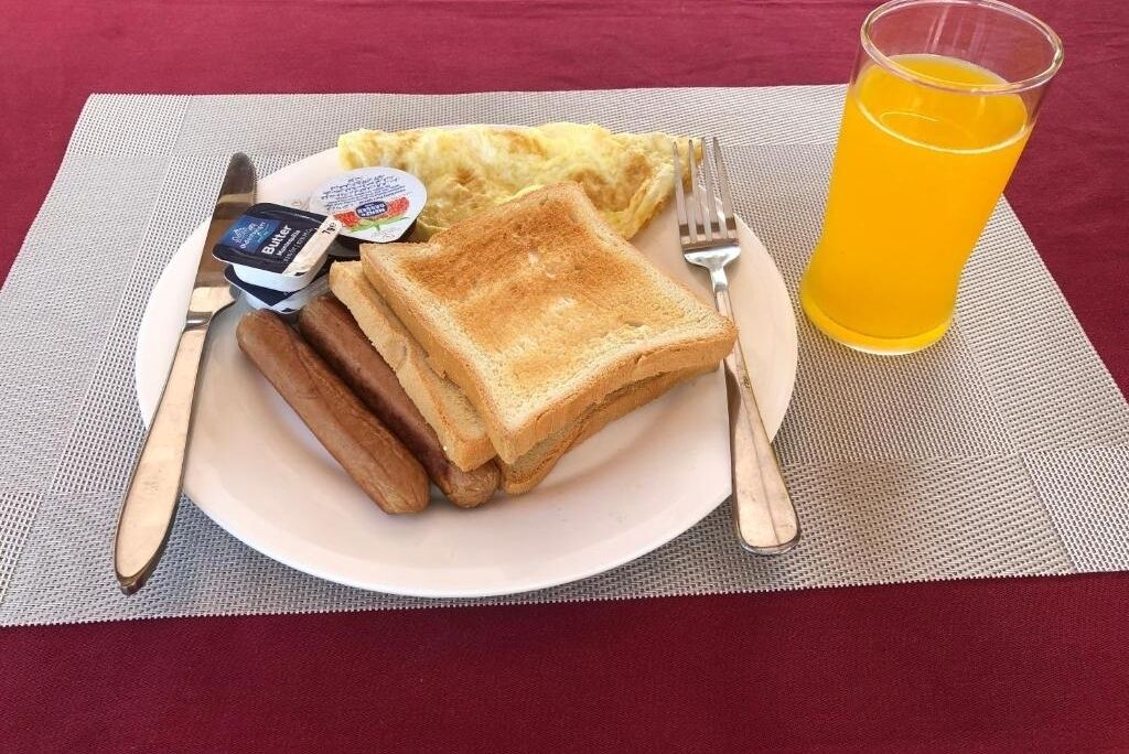 Une assiette de petit-déjeuner avec des toasts, des saucisses, du beurre, de la confiture et un verre de jus d’orange, le tout posé sur une table recouverte d’un set de table.