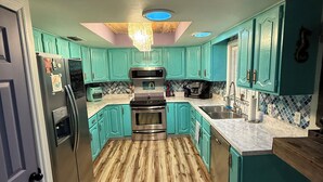 Une cuisine avec des armoires de couleur turquoise, un plancher en bois, un réfrigérateur en acier inoxydable et un lustre.