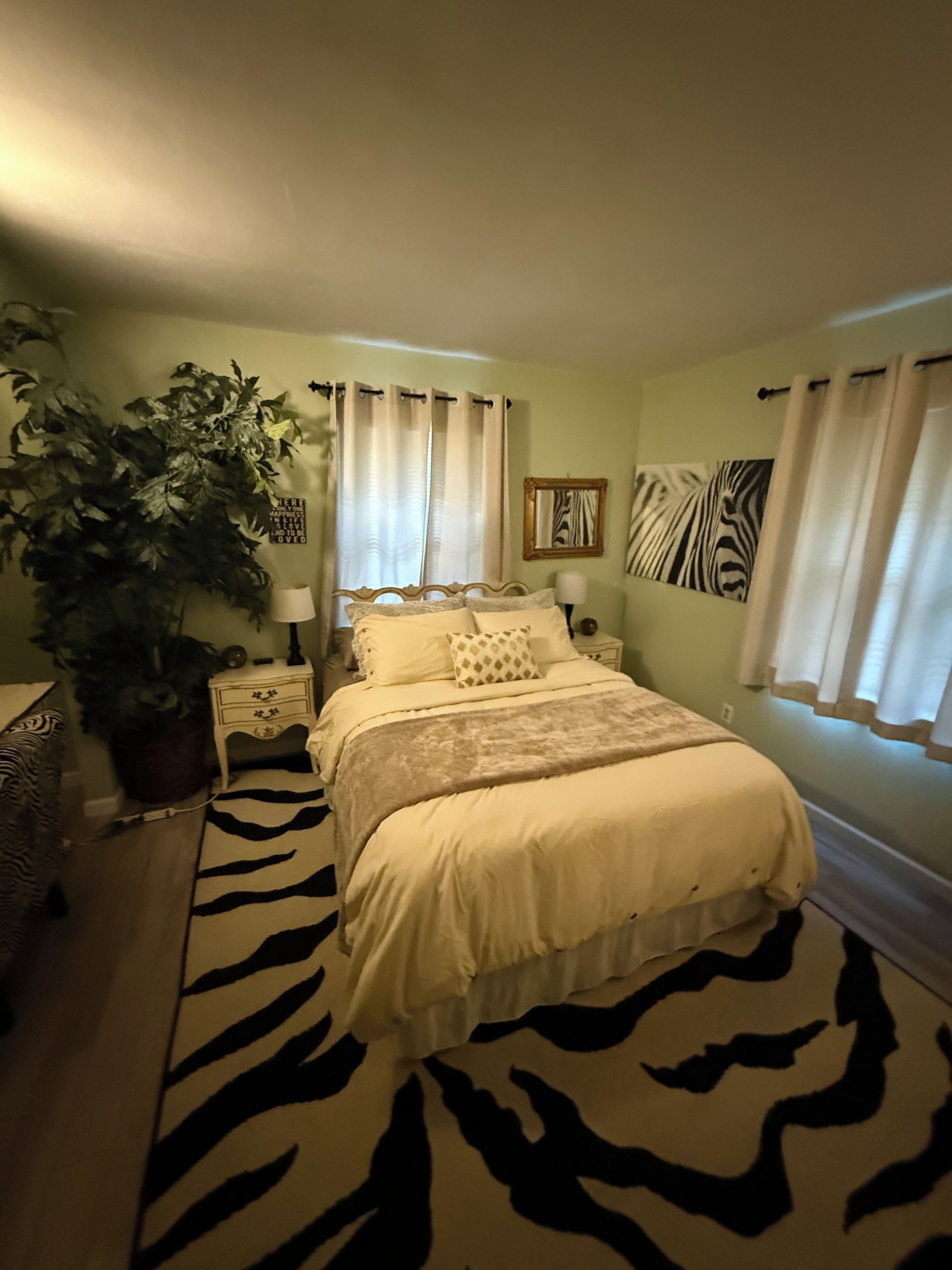 Une chambre avec un tapis à motifs zébrés, une grande plante en pot, une œuvre d’art sur le thème des zèbres et un lit avec une literie blanche.