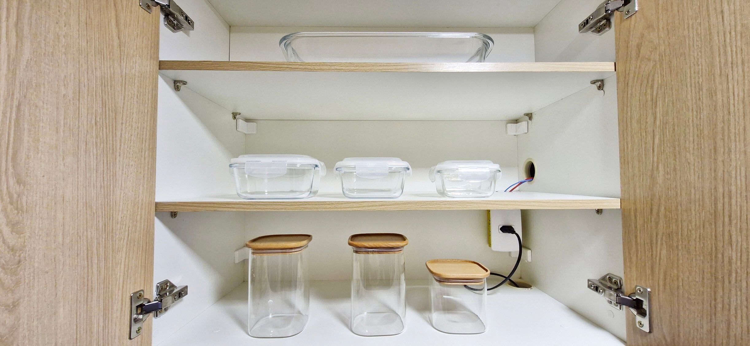 Et køkkenskab med tre klare plastbeholdere på øverste hylde og tre glasbeholdere med trælåg på nederste hylde.