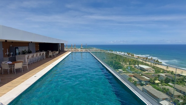 A long swimming pool with a view of the ocean and beach on one side, and a bar area with seating on the other.