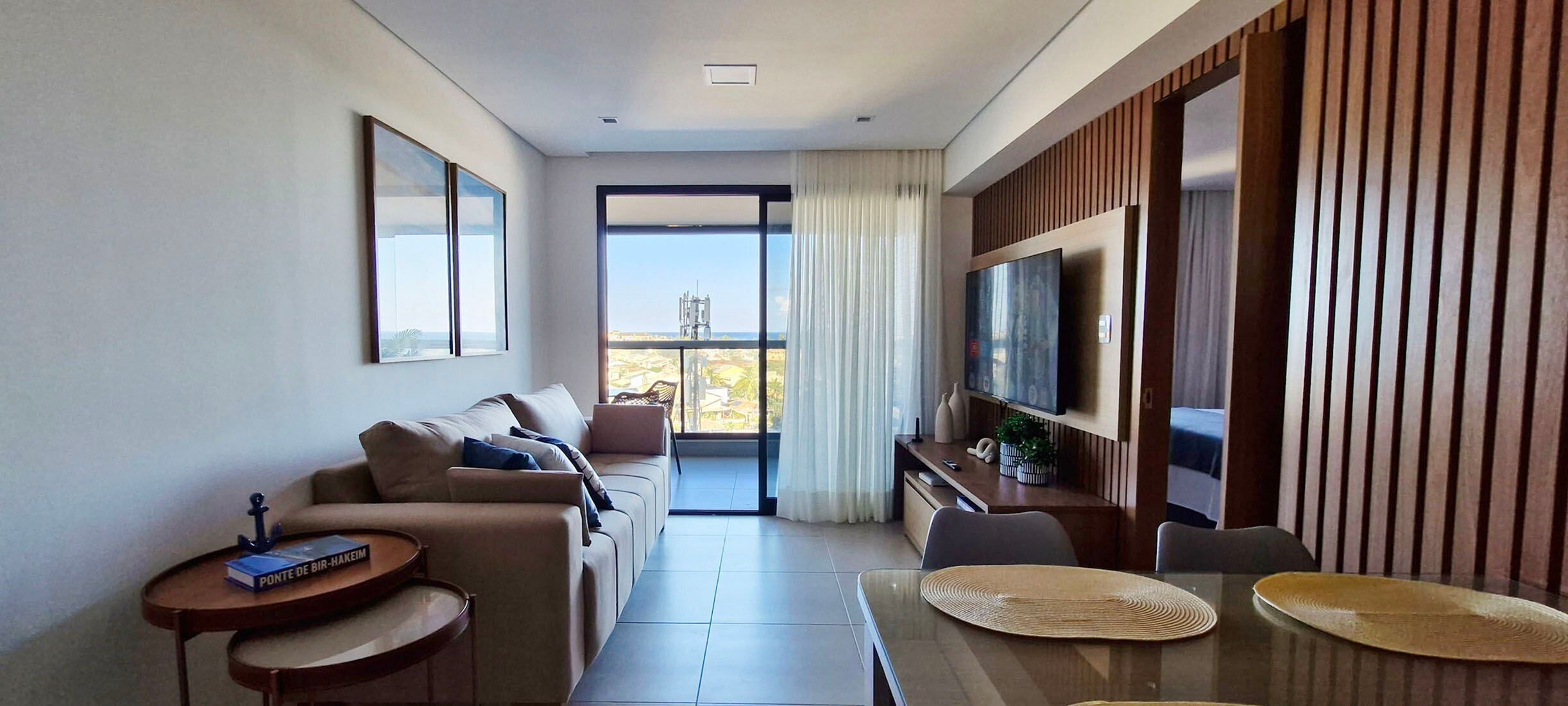A modern hotel room with a sofa, a dining table, a TV, and a balcony with a view.