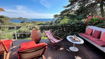 Une terrasse en bois avec des meubles d’extérieur, un grand vase et une vue panoramique sur la mer et les collines.