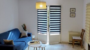 Un salon avec un canapé bleu, une petite table ronde, une lampe suspendue, une porte coulissante avec des stores et un radiateur.