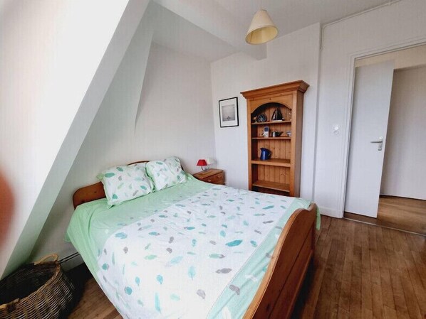 A bedroom with a sloped ceiling, a bed with a patterned bedspread, a wooden wardrobe, and a door to another room.