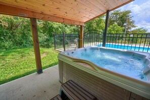 A hot tub under a wooden pergola with a view of a swimming pool and greenery.