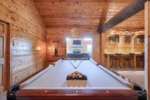 A pool table in a wooden cabin with a stone fireplace and bar seating area.