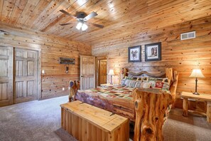 A log cabin bedroom with a bed, a bench, a ceiling fan, and framed pictures on the wall.