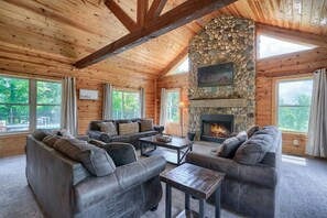 A log cabin interior with a stone fireplace, wooden ceiling beams, and large windows.