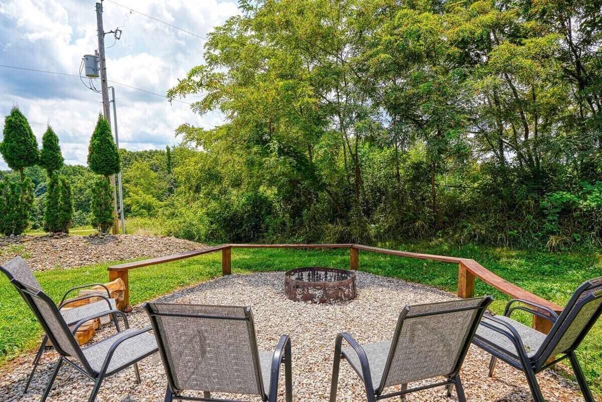 A circular outdoor seating area with a fire pit, surrounded by a wooden fence, set in a lush green landscape with trees and a power line in the background.