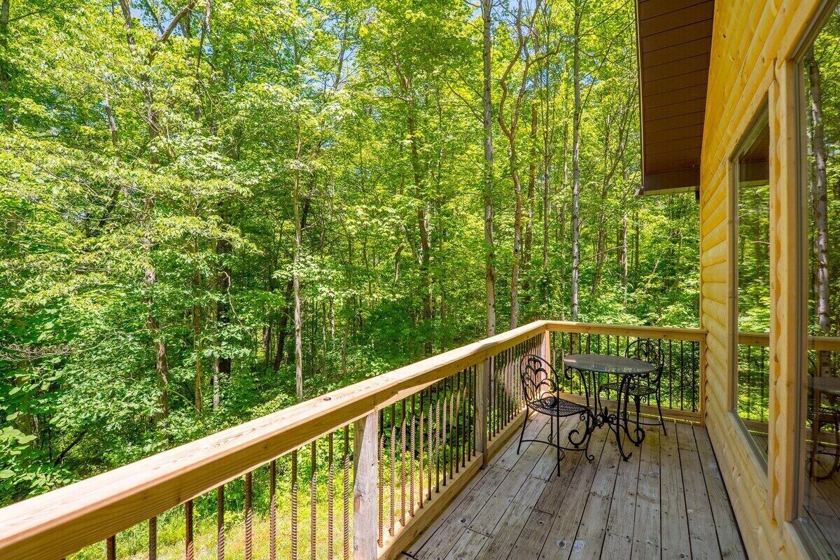 A wooden deck with a metal railing, a table, and chairs, overlooking a dense forest.