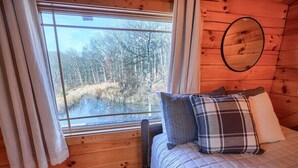 A wooden cabin room with a bed, a window with curtains, and a mirror on the wall.