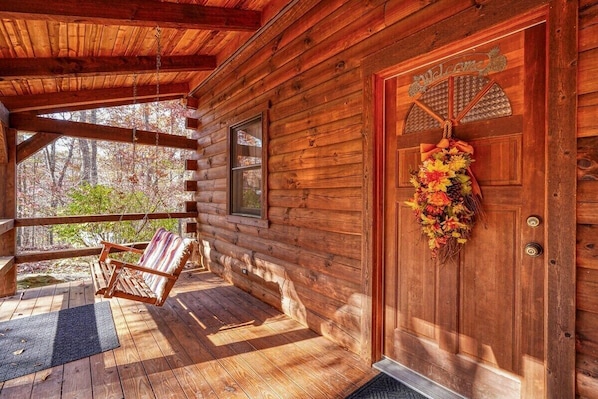 A wooden cabin with a porch, a swing chair, and a door with a 'Welcome' sign.