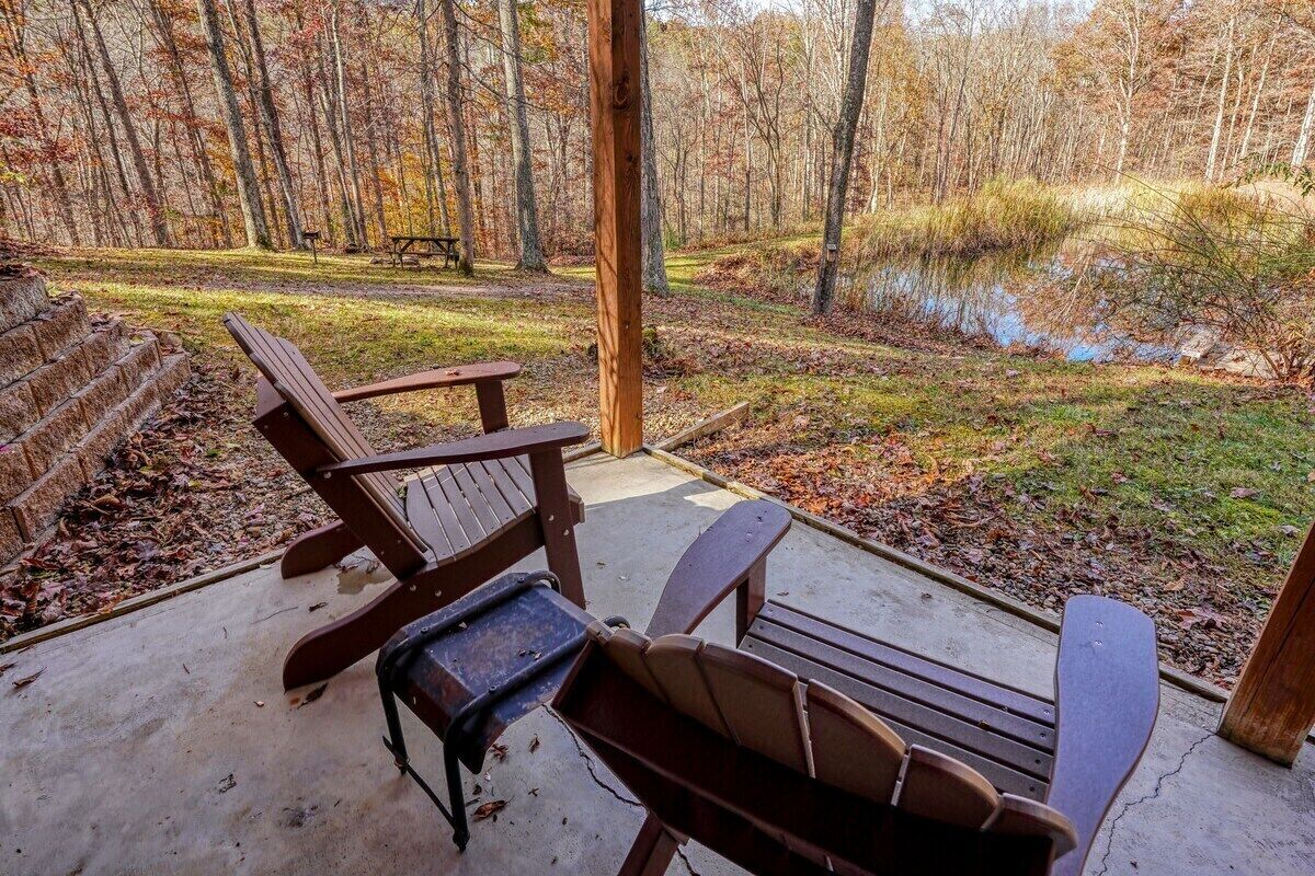 A covered outdoor seating area with Adirondack chairs and a picnic table, overlooking a pond and forested area.