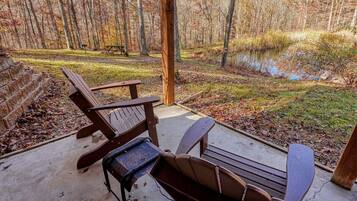 A covered outdoor seating area with Adirondack chairs and a picnic table, overlooking a pond and forested area.