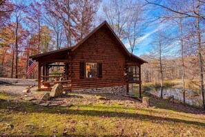 A log cabin with a porch, a stone foundation, and a pond in the background.