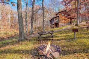 A cabin with a fire pit in the foreground, surrounded by trees.