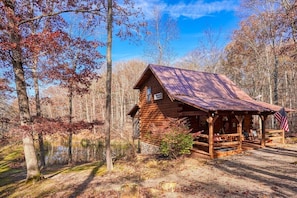 A rustic cabin with a porch and a small deck, surrounded by trees and a body of water.