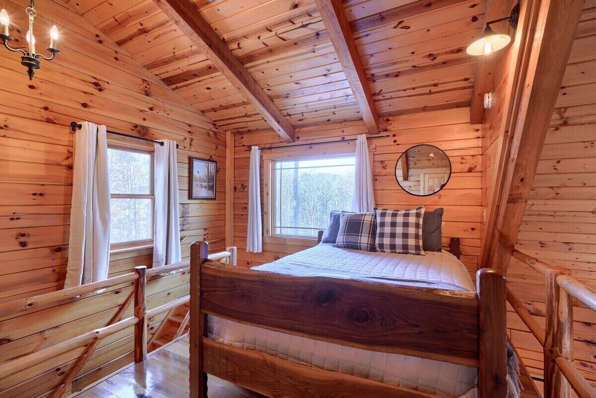 Ein Schlafzimmer einer Holzhütte mit einem Bett, zwei Fenstern, einer Chandelieber und einem Spiegel an der Wand.