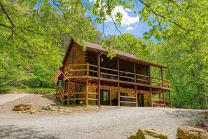 A rustic log cabin with a porch and a stone foundation, surrounded by lush greenery.