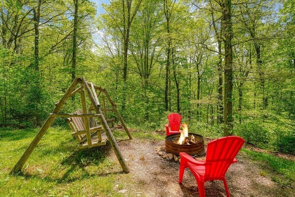 Un décor forestier avec une balançoire en bois, un foyer et des chaises rouges.