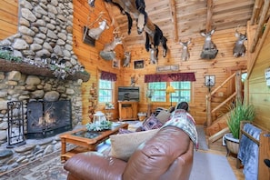A log cabin interior with a stone fireplace, wooden walls, and deer antlers mounted on the ceiling.