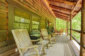 A wooden cabin with a covered porch, furnished with wooden chairs and a table, and surrounded by greenery.
