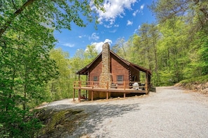 A rustic cabin with a stone chimney, wooden deck, and a gravel driveway surrounded by forest.