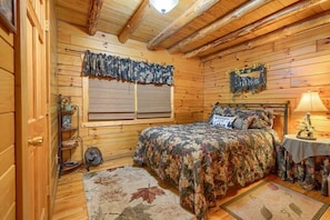 A cozy wooden cabin bedroom with a bed, a small table with a lamp, a window with a curtain, and a shelf with decorative items.