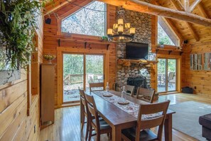 A wooden cabin with a stone fireplace, a dining table with chairs, and large windows.