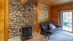 A cozy cabin interior with a stone wall, a black wood-burning stove, and a cushioned sofa by a large window.