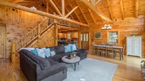 A log cabin interior with a large wooden staircase, a sectional sofa, a dining area, and a kitchen.