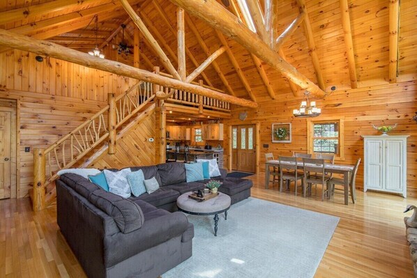 A log cabin interior with a large wooden staircase, a sectional sofa, a dining area, and a kitchen.