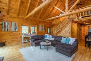 A log cabin interior with a large wooden staircase, a sectional sofa, and a dining area.