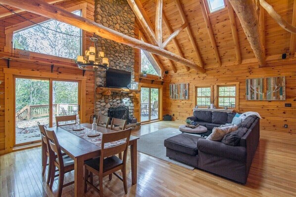 A spacious log cabin with a stone fireplace, wooden dining table, and a cozy living area with a sofa and TV.