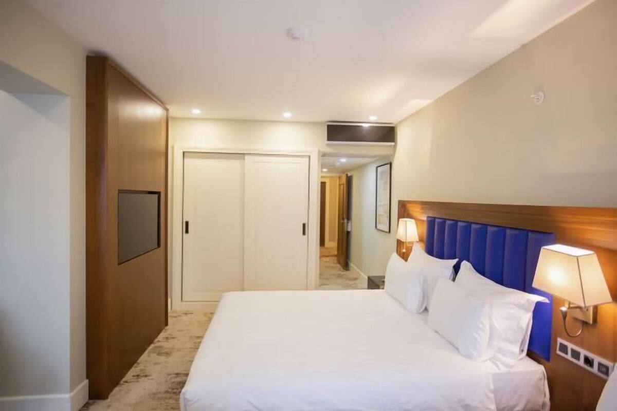 Une chambre d’hôtel avec un lit, des lampes de chevet, une tête de lit en bois, une porte et un couloir.