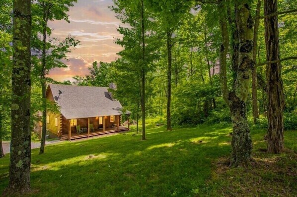 A log cabin with a porch, surrounded by a forest.