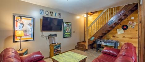 Uma sala de estar aconchegante com sofás de couro vermelho, uma televisão de tela plana, pôsteres emoldurados e um lance de escadas de madeira.