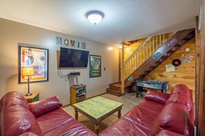 A cozy living room with red leather sofas, a flat-screen TV, framed posters, and a wooden staircase.