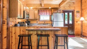 A wooden cabin kitchen with a central island, bar stools, a refrigerator, and a window with a plaid curtain.