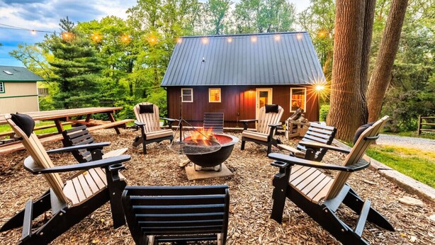 A wooden cabin with a fire pit in the center, surrounded by Adirondack chairs.