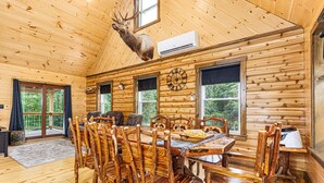 A wooden cabin with a dining area featuring a large wooden table and chairs, a mounted deer head, and a clock on the wall.